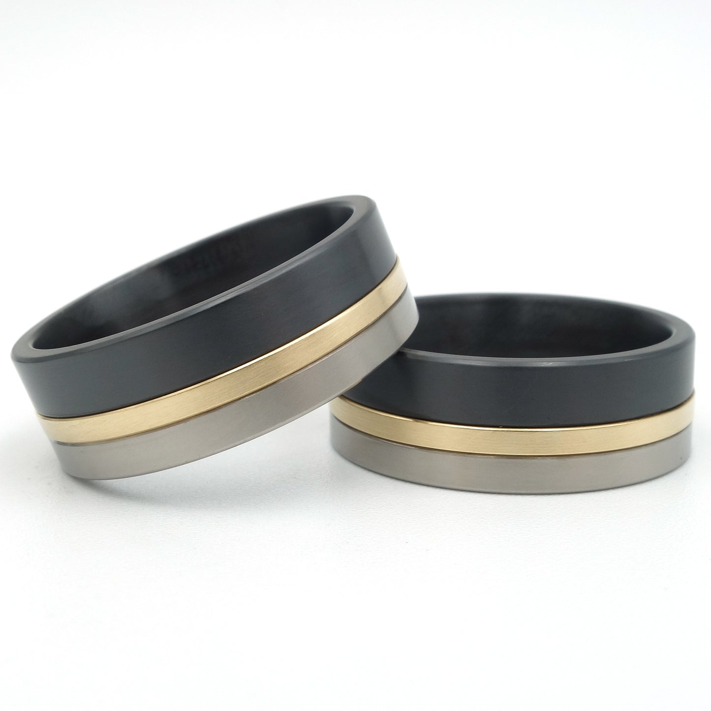 Twee ringen in titanium, geelgoud en zwart zirkonium, 8,3mm breed, met een smalle gouden lijn tussen grijs titanium en zwart zirkonium.