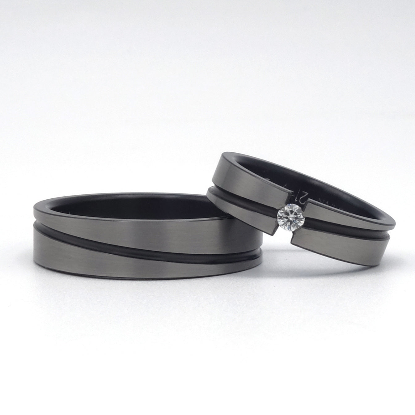 Twee zirkoniumringen met diagonale zwarte lijn; één ring van 5 mm met diamant in semi-spanzetting, één van 6 mm zonder steen, mat afgewerkt met zichtbaar kaal zirkonium.