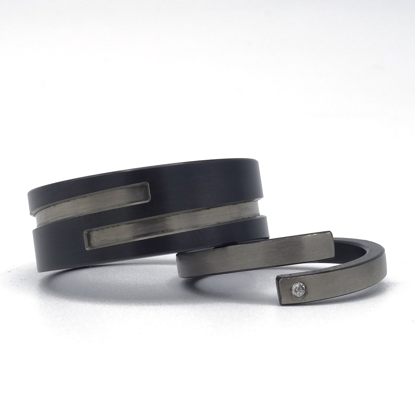 Set moderne trouwringen in zirkonium met omgekeerde kleuren: brede zwarte ring met grijze inleg en smalle grijze ring met zwarte binnenzijde en diamantje.