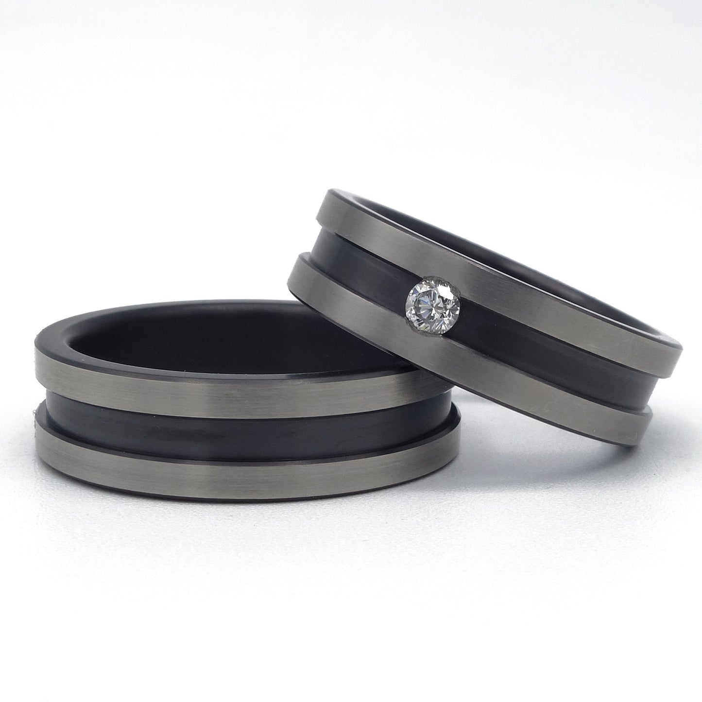 Twee zirkonium ringen met brede zwarte groef; één ring met diamant van 0.08 ct en één zonder, op een witte achtergrond.