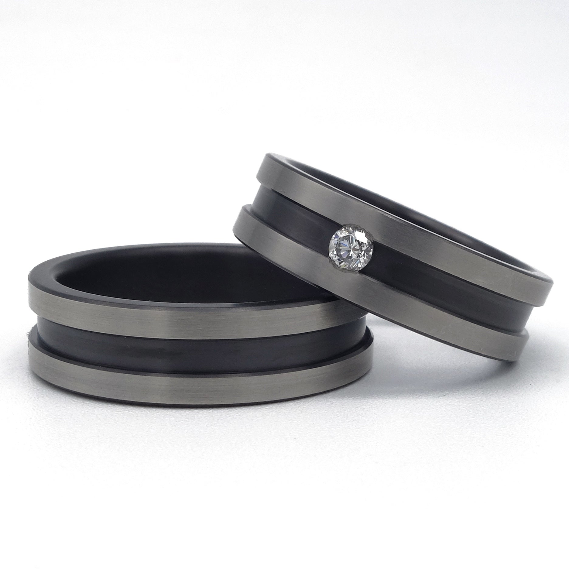 Twee zirkonium ringen met brede zwarte groef; één ring met diamant van 0.08 ct en één zonder, op een witte achtergrond.