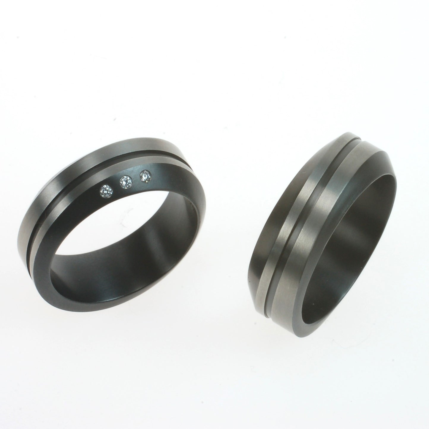 Set van twee zirkonium trouwringen met golvend profiel en zwarte lijn; één ring met drie diamanten, één zonder.