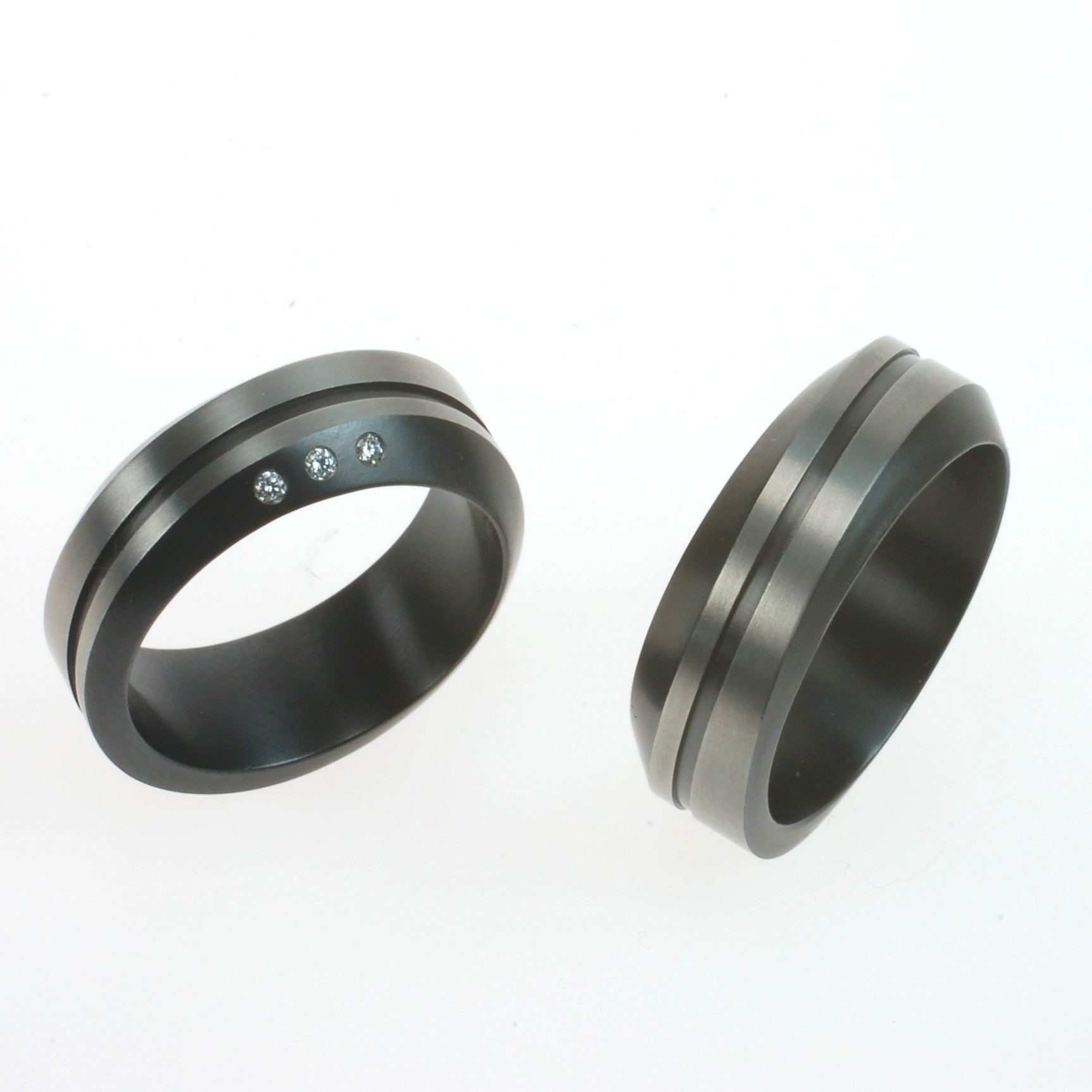 Set van twee zirkonium trouwringen met golvend profiel en zwarte lijn; één ring met drie diamanten, één zonder.