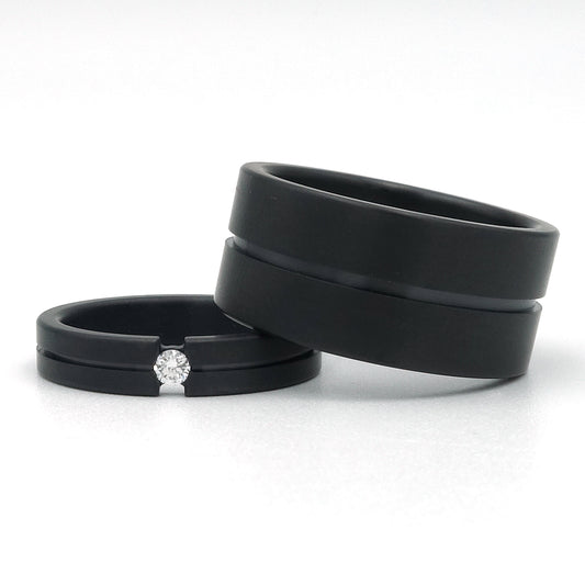 Twee zwarte zirkoniumringen; één van 4 mm met diamant in semi-spanzetting, en één brede ring van 10 mm zonder steen, beide met matte buitenkant en glanzende lijn.
