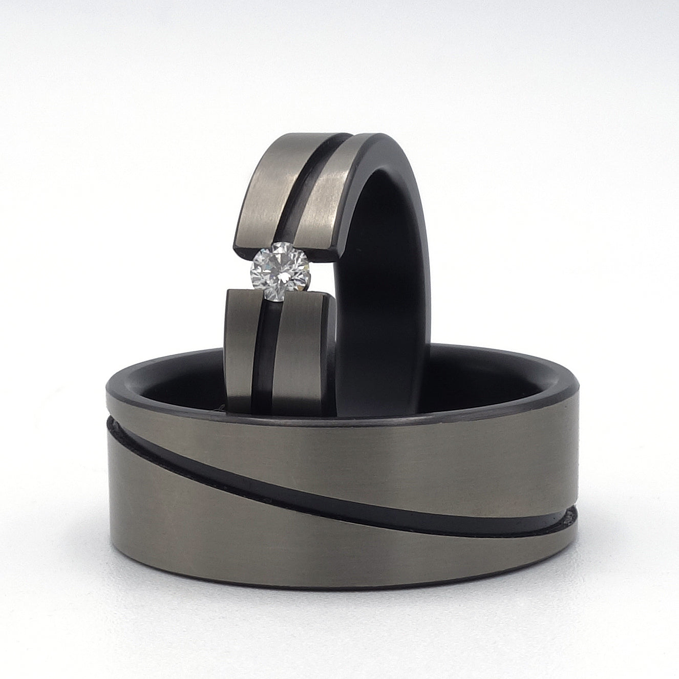 Twee zirkoniumringen met diagonale matte groef; damesring met diamant in semi-spanzetting, herenring zonder steen. Contrasterend mat en kaal zirkonium.