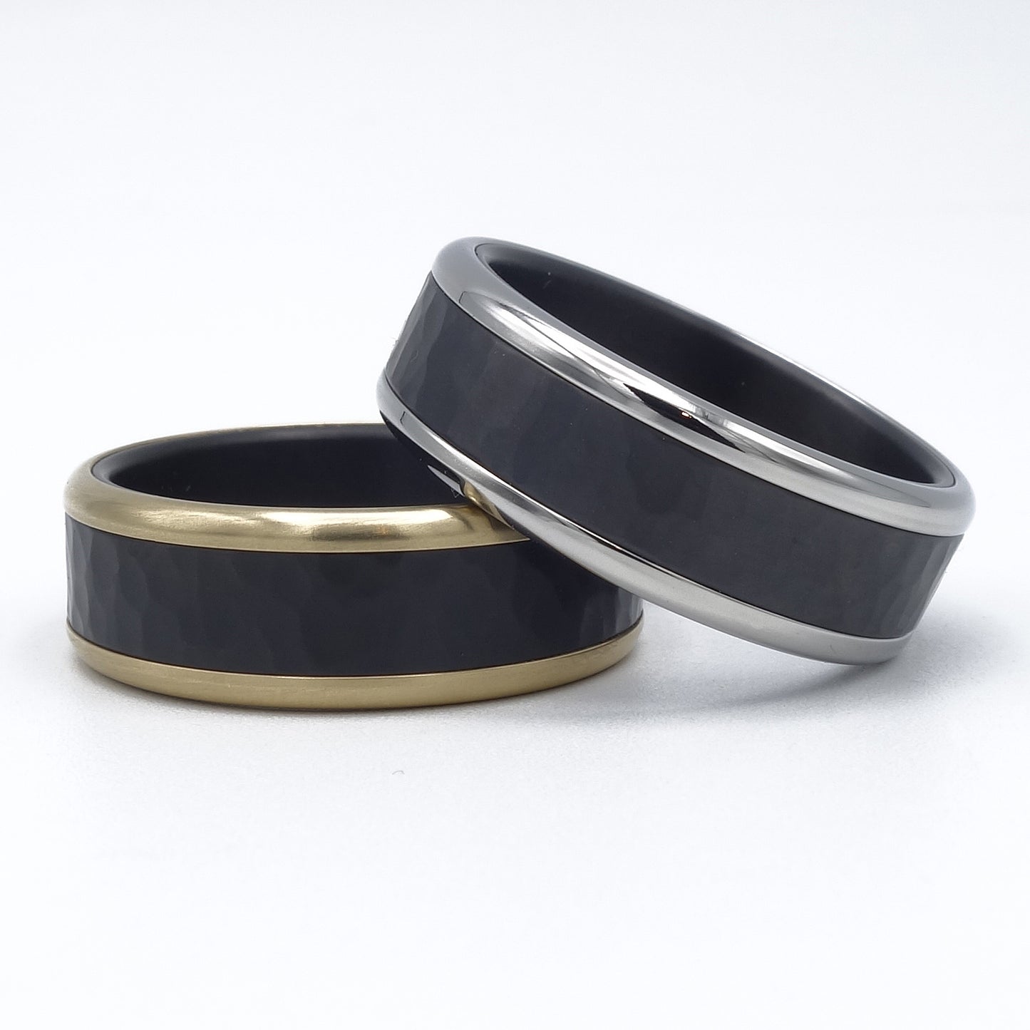 Twee ringen van zwart zirkonium met hamerslag. Damesring met geelgouden rand, herenring met glanzend edelstalen rand. Moderne, luxe trouwringen.