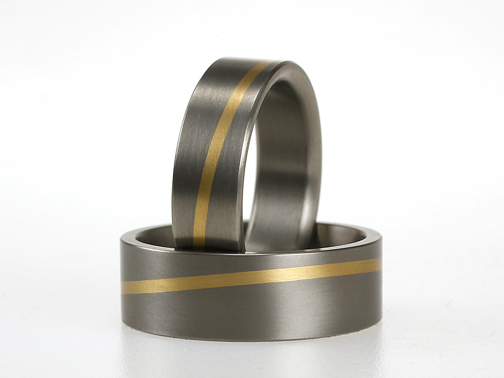Twee titanium ringen met diagonale inleg van 18 karaat geelgoud; een smallere en een bredere variant, op een witte achtergrond.