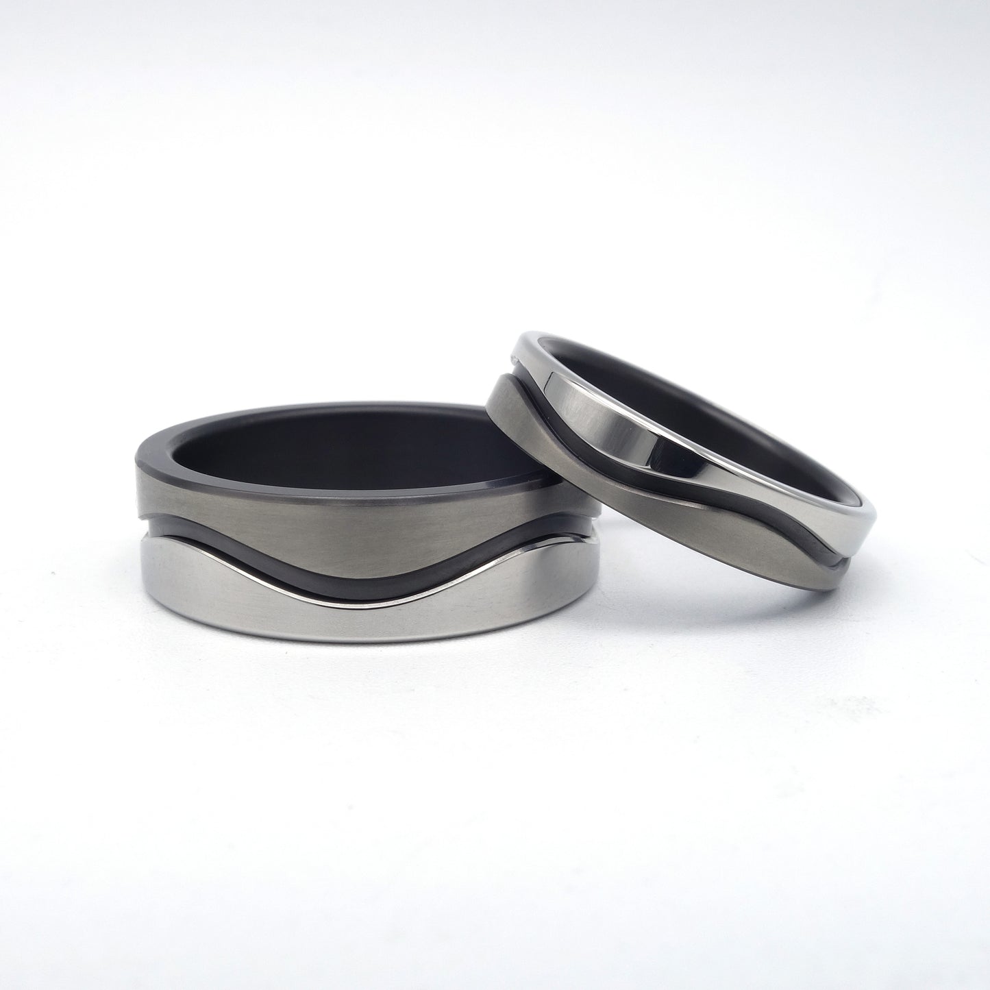 Twee trouwringen uit de Wave-serie van zwart zirkonium en edelstaal met golvend ontwerp, in breedtes van 4 en 7 mm, samen gepresenteerd.