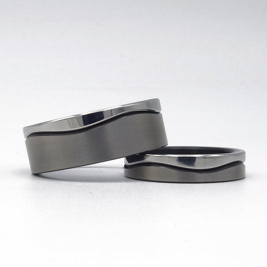 Twee trouwringen uit de Wave Fijn-serie van zwart zirkonium en edelstaal met smalle golvende lijn, gepresenteerd op een witte achtergrond.