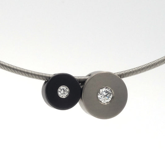 Twee ronde hangers aan één ketting: zwarte zirkonium schijf met 0,03 ct diamant en titanium schijf met 0,08 ct diamant.