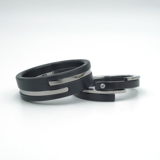 Twee ringen met modern design: brede ring in zwart zirkonium met edelstalen inleg, smalle ring met diamant. Symbolische set trouwringen.