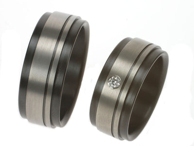 Twee zirkonium trouwringen met matte middenbaan en zwarte randen. Eén ring met diamant, strak en modern ontwerp.