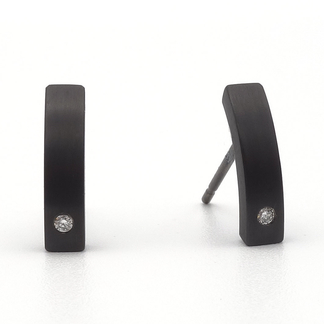 Zwarte titanium oorstekers met een subtiele kromming en een kleine briljant geslepen diamant aan de onderzijde, gefotografeerd op een witte achtergrond.