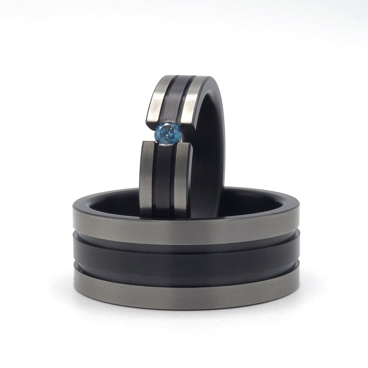 Twee zirkoniumringen met zwarte lijnen; één met blauwe diamant in semi-spanzetting en één bredere ring zonder steen, op een witte achtergrond.