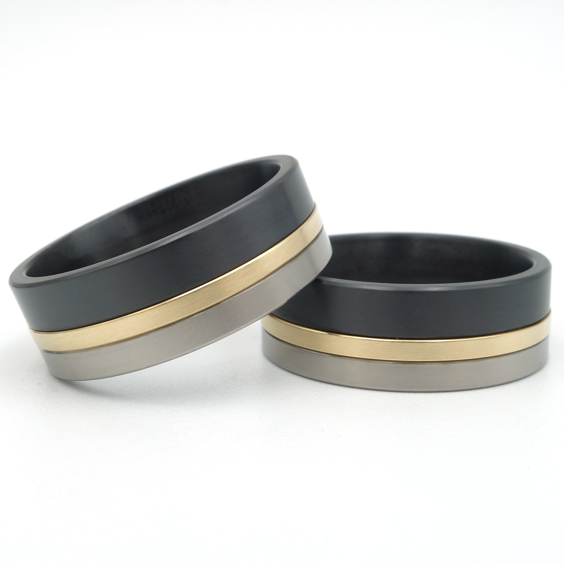 Twee ringen in titanium, geelgoud en zwart zirkonium, 8,3mm breed, met een smalle gouden lijn tussen grijs titanium en zwart zirkonium.