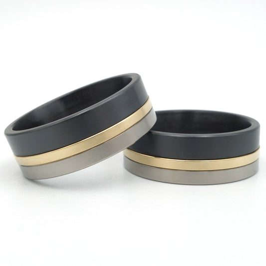 Twee ringen in titanium, geelgoud en zwart zirkonium, 8,3mm breed, met een smalle gouden lijn tussen grijs titanium en zwart zirkonium.