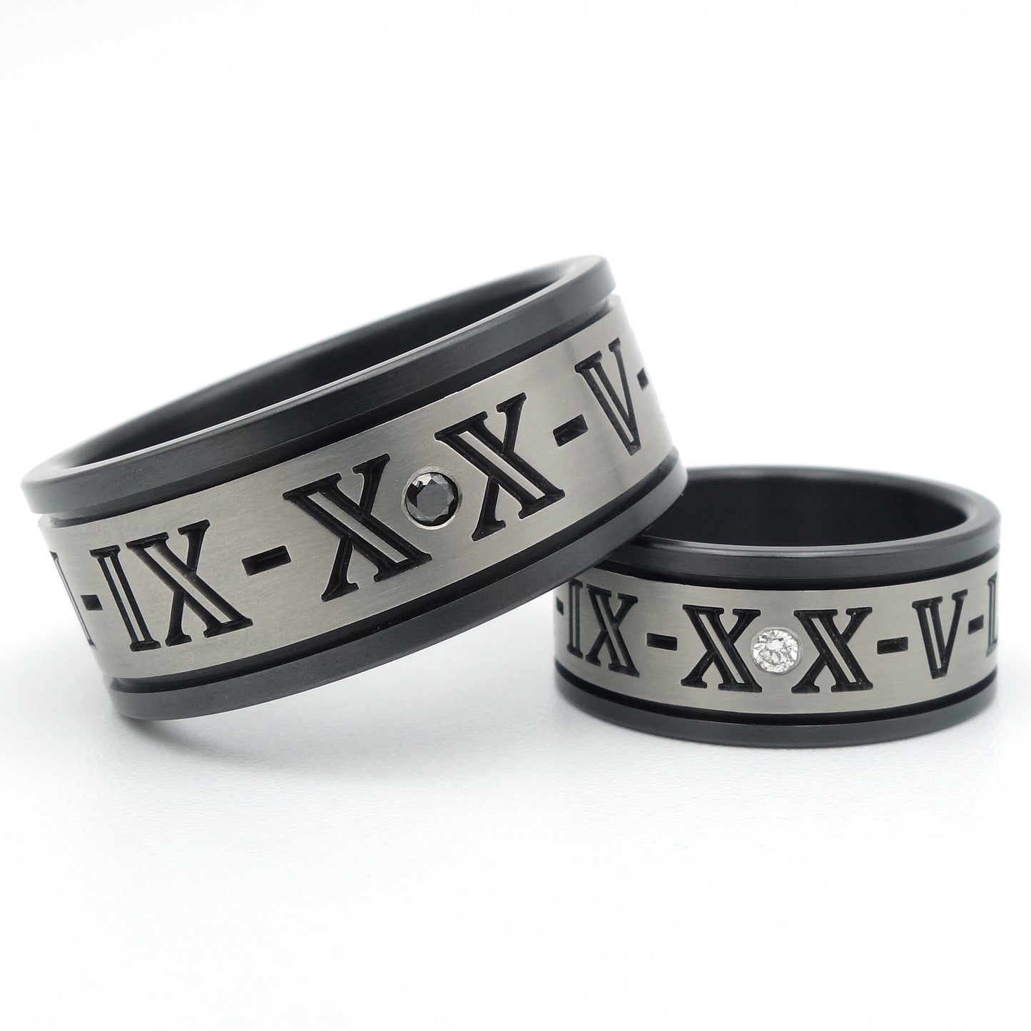 Twee zirkonium ringen met gegraveerde Romeinse cijfers, grijs middengedeelte met zwart geoxideerde randen, één met zwarte diamant en één met witte diamant als accent.