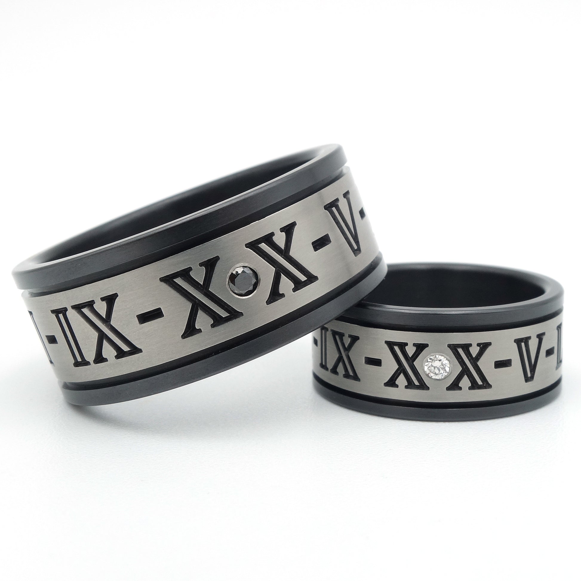 Twee zirkonium ringen met gegraveerde Romeinse cijfers, grijs middengedeelte met zwart geoxideerde randen, één met zwarte diamant en één met witte diamant als accent.