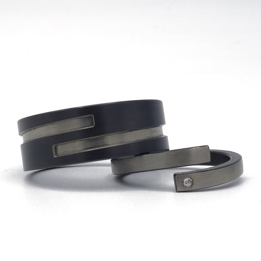 Set moderne trouwringen in zirkonium met omgekeerde kleuren: brede zwarte ring met grijze inleg en smalle grijze ring met zwarte binnenzijde en diamantje.