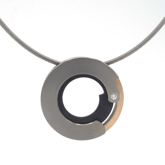 Moderne ronde hanger van titanium en roodgoud met zwarte inleg en diamant, aan een strakke slangenketting.

