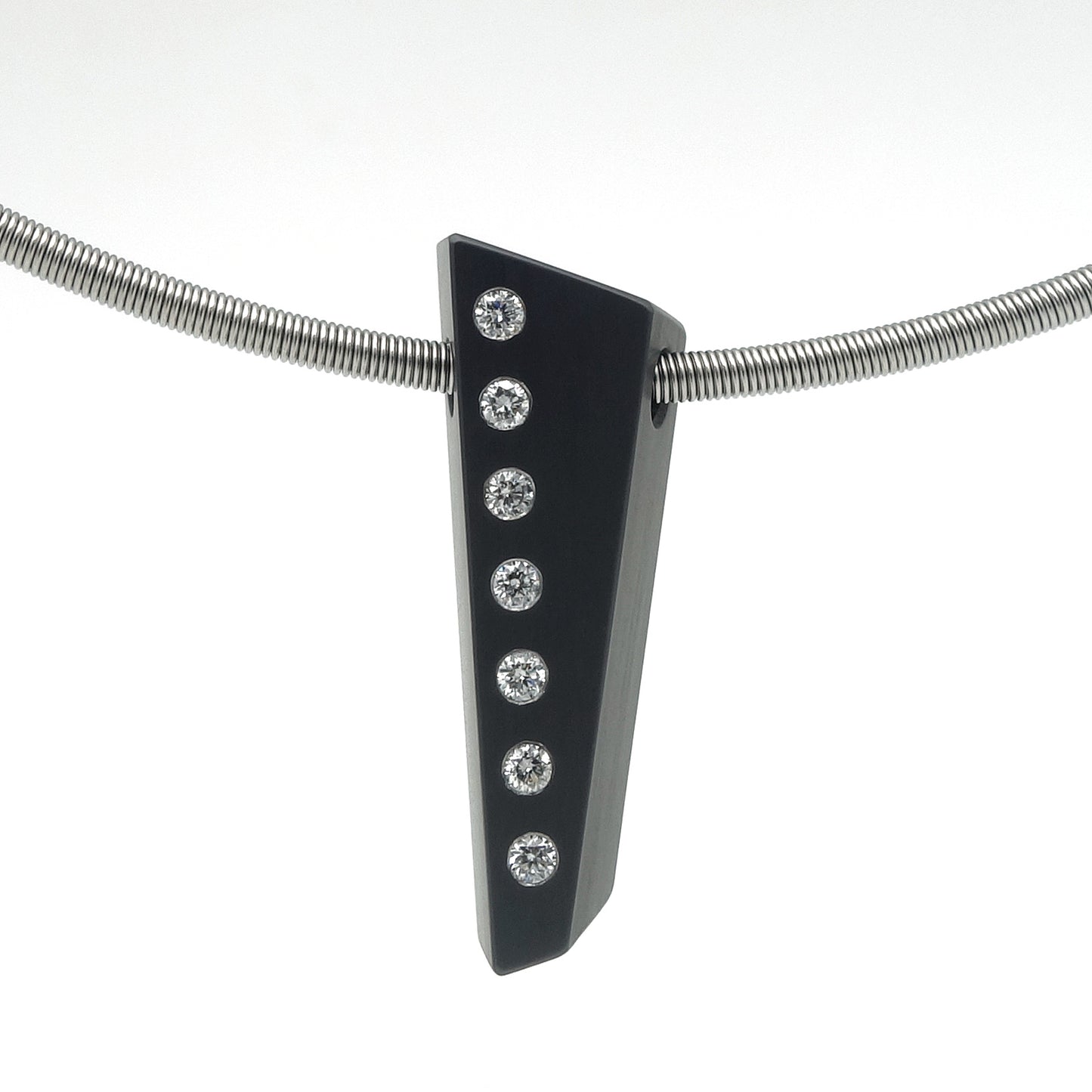Asymmetrische hanger van zwart zirkonium met zeven flush ingezette briljant geslepen diamanten in een verticale lijn, hangend aan een RVS omega-spang, op witte achtergrond.