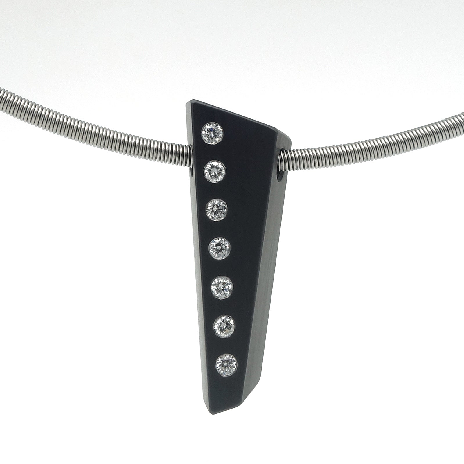 Asymmetrische hanger van zwart zirkonium met zeven flush ingezette briljant geslepen diamanten in een verticale lijn, hangend aan een RVS omega-spang, op witte achtergrond.