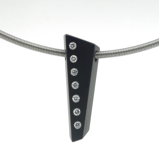 Asymmetrische hanger van zwart zirkonium met zeven flush ingezette briljant geslepen diamanten in een verticale lijn, hangend aan een RVS omega-spang, op witte achtergrond.