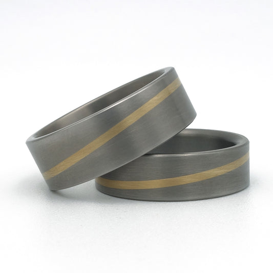 Twee titanium ringen met diagonale inleg van 18 karaat geelgoud; een smallere en een bredere variant, op een witte achtergrond.