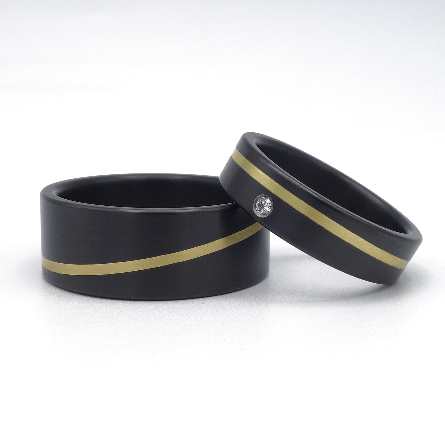 Twee zwarte zirkonium ringen et een diagonale lijn van geelgoud,. De ene ring is breder dan de andere. De smalle ring heeft een diamantje erin.