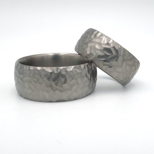 Brede titanium trouwringen met grof hamerslagpatroon; damesring 8 mm, herenring 10 mm.