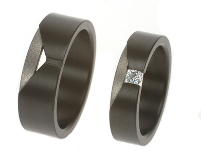Twee moderne ringen in zwart en grijs met V-vormige uitsparing, één met witte diamant. Strakke trouwringen van titanium en zirkonium.