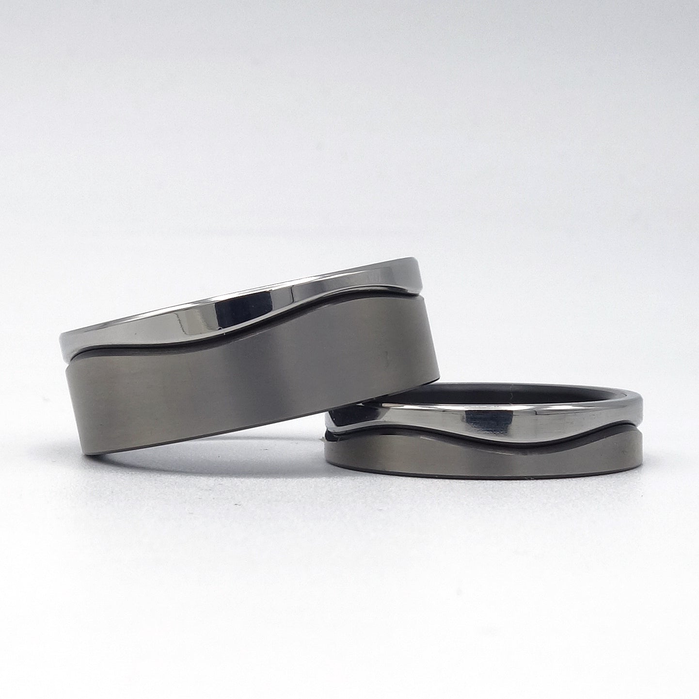 Twee trouwringen uit de Wave Fijn-serie van zwart zirkonium en edelstaal met smalle golvende lijn, gepresenteerd op een witte achtergrond.