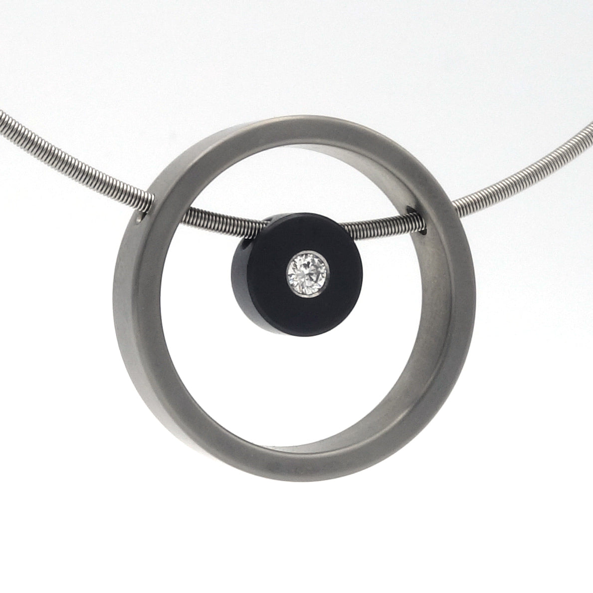 Moderne hanger met zwarte zirkonium schijf en 0,08 ct diamant, geplaatst in een ronde titanium ring aan een slangenketting.

