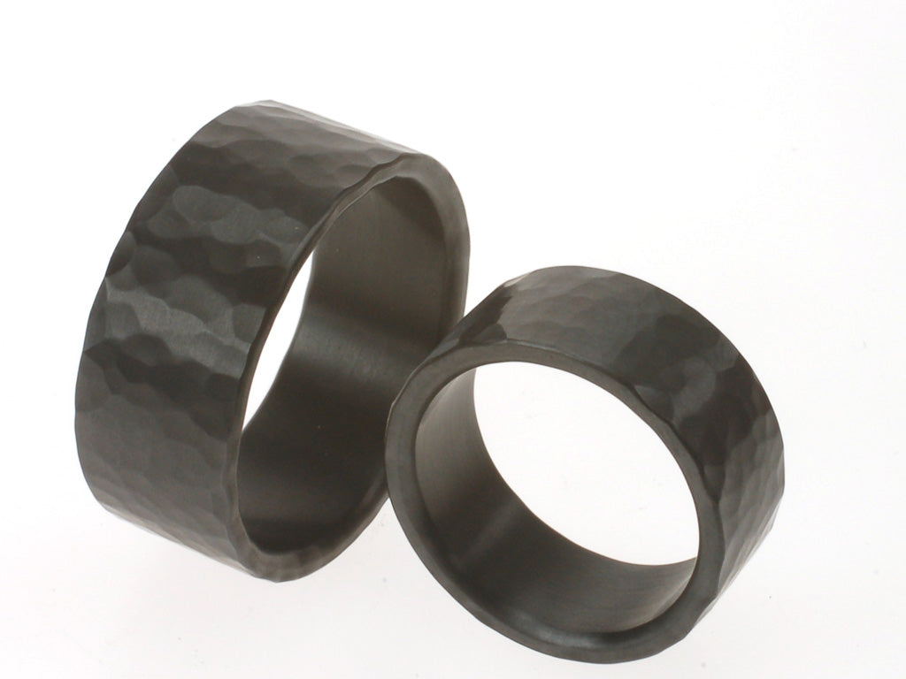 Twee brede zwarte ringen van zirkonium met grove hamerslagstructuur. Stoere, matte trouwringen met unieke textuur.