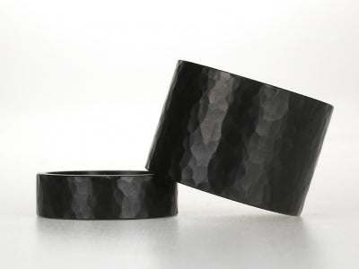 Twee brede zwarte zirkoniumringen met grof hamerslagpatroon, een smalle en een extra brede variant.