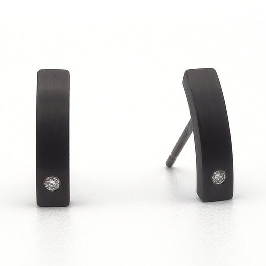 Zwarte titanium oorstekers met een subtiele kromming en een kleine briljant geslepen diamant aan de onderzijde, gefotografeerd op een witte achtergrond.
