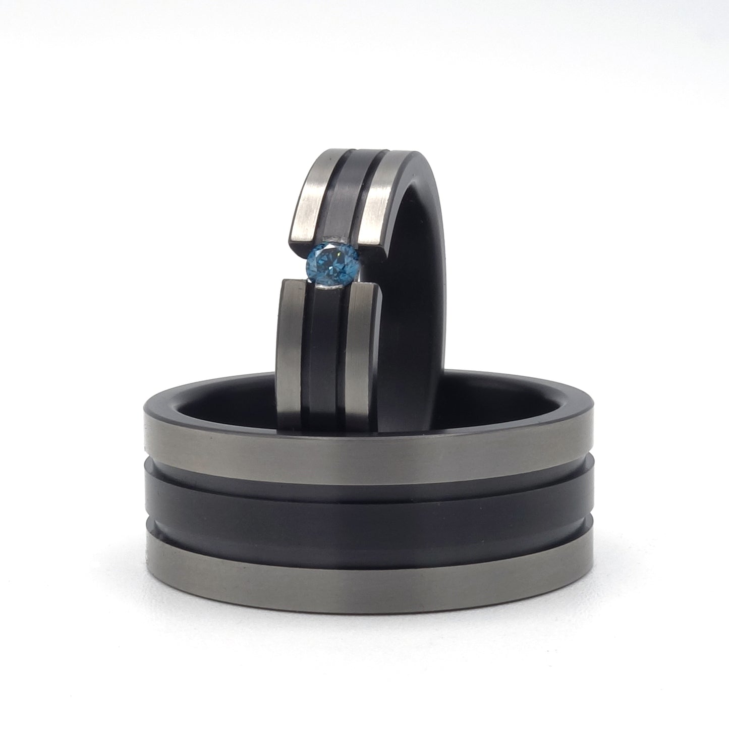 Twee zirkoniumringen met zwarte lijnen; één met blauwe diamant in semi-spanzetting en één bredere ring zonder steen, op een witte achtergrond.